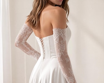 May include: A white strapless gown featuring a corset-style bodice with a lace-up back closure and detachable long sheer lace sleeves with floral patterns. The skirt is made of smooth fabric and flows downward from the waistline.