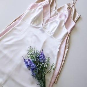 May include: Three silky slip dresses in ivory, blush pink, and champagne gold, layered on top of each other. A bouquet of purple and white flowers is in the center of the image.