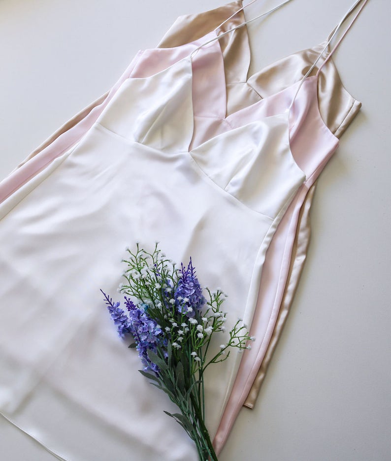 May include: Three silky slip dresses in ivory, blush pink, and champagne gold, layered on top of each other. A bouquet of purple and white flowers is in the center of the image.
