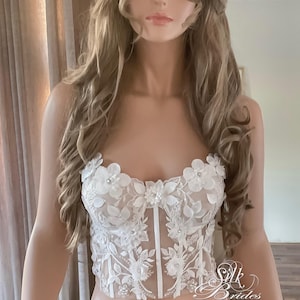 May include: White lace bridal corset top with floral appliques and a sheer bodice. The corset has a sweetheart neckline and is adorned with delicate beading.