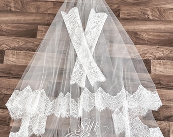 Op de afbeelding: Een witte bruidssluier met ingewikkelde kanten details langs de randen en op de bijgevoegde handschoenen. De sluier is doorschijnend en het kant heeft een delicaat bloemenpatroon. De woorden "Silk Brides" zijn zichtbaar.