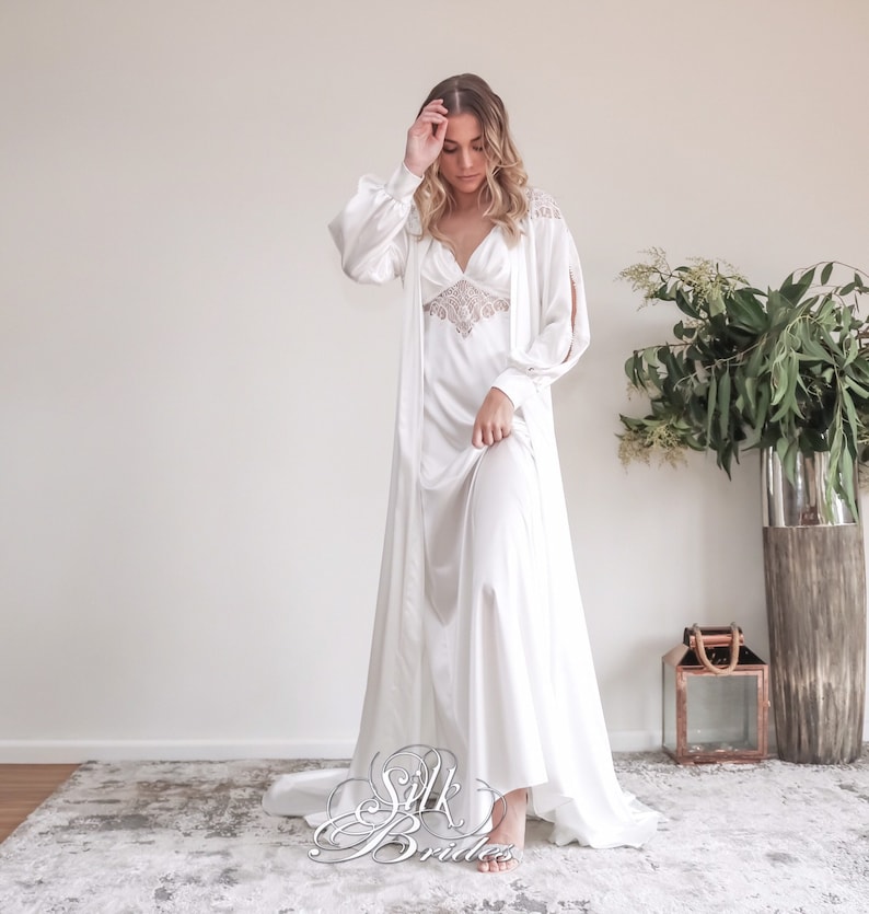 May include: A white silk robe with long sleeves and a V-neckline. The robe has a lace trim around the neckline and a satin finish. The model is wearing the robe with the sleeves rolled up and is standing on a rug.