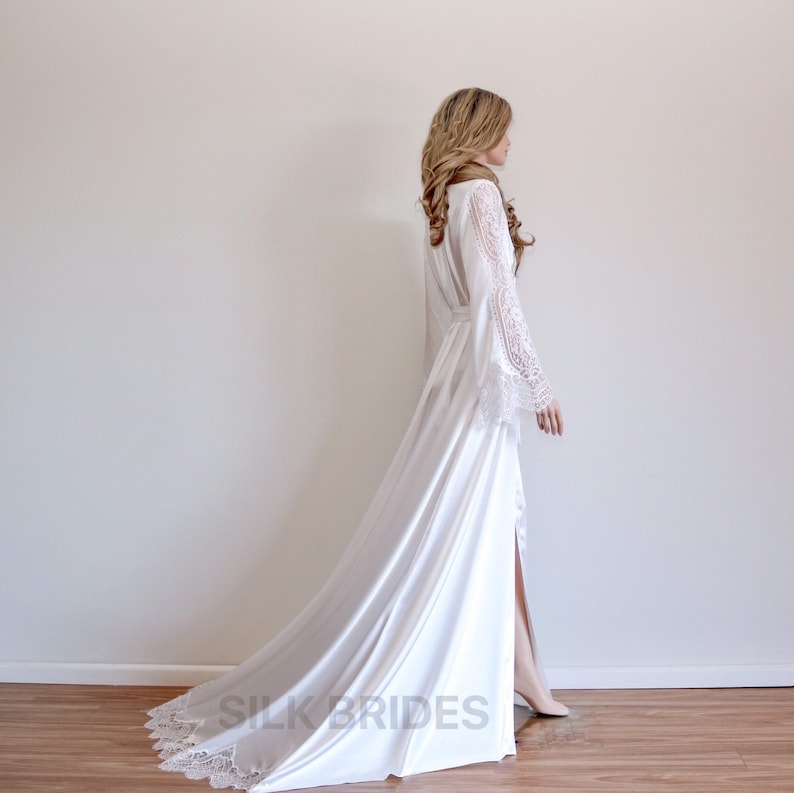 May include: A white satin robe with long sleeves and a lace trim. The robe has a long train and a slit up the front. The model is wearing the robe and standing on a wooden floor.