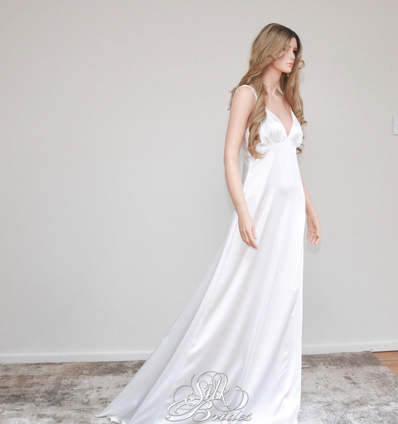 May include: A white silk wedding dress with a simple, elegant design. The dress has a V-neckline and spaghetti straps. The skirt is flowing and has a slight train.
