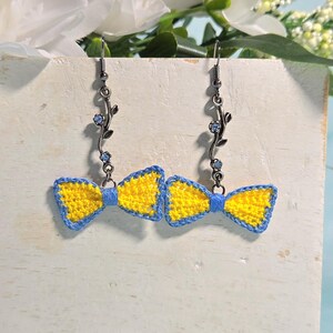 Könnte beinhalten: Handgefertigte Ohrringe mit gelben und blauen gehäkelten Fliegen. Die Schleifen sind blau umrandet und hängen an dekorativen silberfarbenen Haken mit floralen und blauen Kristallakzenten. Präsentiert vor einem neutralen Hintergrund.