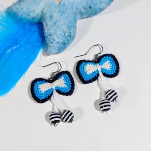May include: Handmade earrings with a bow tie design in blue, white, and black, with striped beads hanging below. The earrings are presented on a white surface, with a blue starfish and feather in the background.