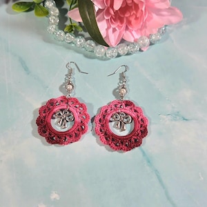May include: A pair of pink crochet earrings. Each earring has a circular design with a silver bow charm. The earrings have silver hooks and a small pearl. The background includes a pink flower and a string of clear beads. The earrings are handmade.