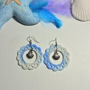 Può includere: Orecchini all'uncinetto fatti a mano con un ciondolo a forma di conchiglia. Gli orecchini presentano un bordo smerlato bianco e azzurro, con un pendente a forma di conchiglia color argento. Sono esposti su una superficie bianca con una stella marina e piume.