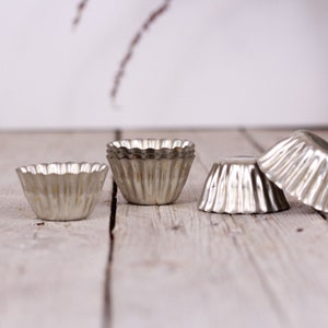 May include: Four silver metal cupcake liners or molds on a light wood surface. The liners are fluted and have a scalloped edge.