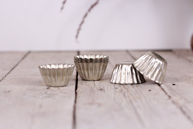 May include: Four silver metal cupcake liners or molds on a light wood surface. The liners are fluted and have a scalloped edge.