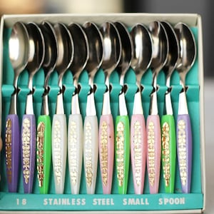 Puede incluir: Conjunto de 18 cucharaditas de acero inoxidable con mangos de plástico de colores. Los mangos tienen un diseño dorado y el texto "18 Stainless Steel Small Spoon" está impreso en la caja.