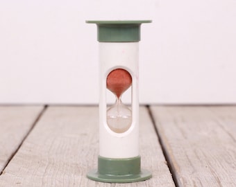 May include: A white hourglass with a green top and base. The hourglass is filled with red sand and is sitting on a wooden surface.