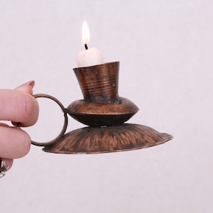 Puede incluir: Un candelero de cobre con un asa de anillo y una sola vela blanca que arde en su interior.