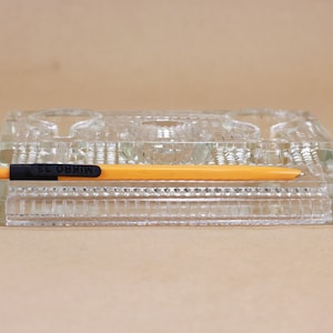 May include: A clear glass pen holder with three compartments and a raised design. A yellow pen with black writing on it is resting on top of the holder.