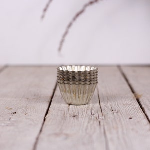 May include: A stack of six silver metal cupcake liners, or baking cups, on a white wooden surface.