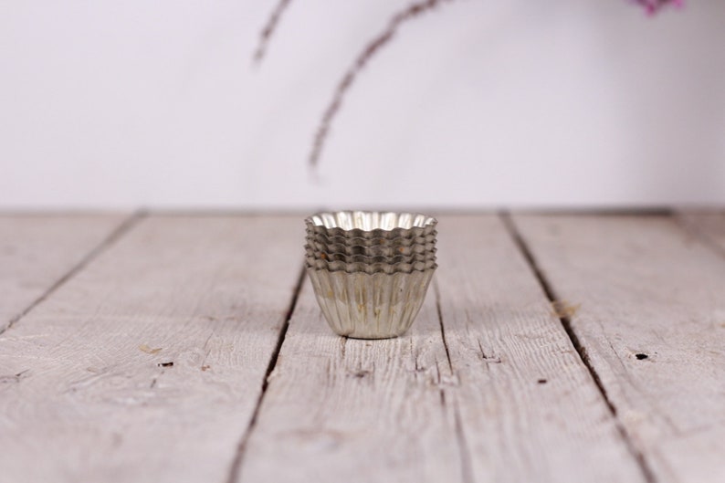 May include: A stack of six silver metal cupcake liners, or baking cups, on a white wooden surface.