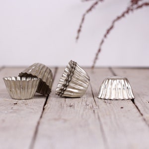 May include: Four silver metal cupcake molds on a white wooden surface. The molds are fluted and have a vintage look.