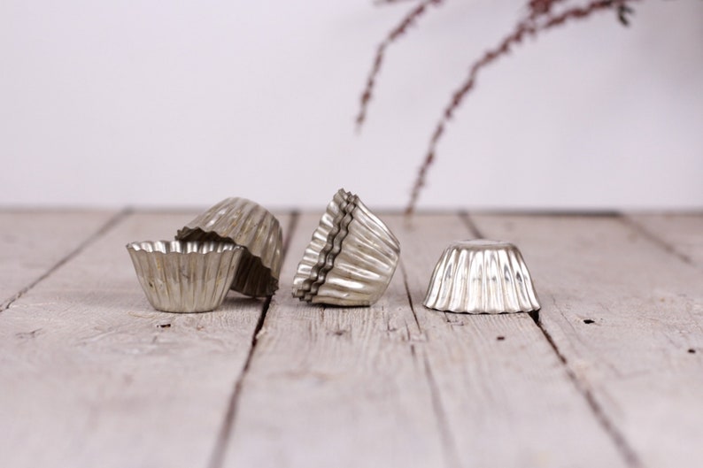 May include: Four silver metal cupcake molds on a white wooden surface. The molds are fluted and have a vintage look.