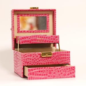 Può includere: Portagioie rosa aperto con una texture a pelle di coccodrillo. La scatola ha uno specchio all'interno del coperchio, un piccolo cassetto e una chiusura color oro. L'interno è beige chiaro.
