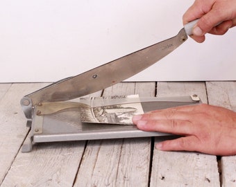 May include: A silver paper cutter with a black handle and a metal blade. The cutter is on a wooden surface and a hand is holding the blade down.