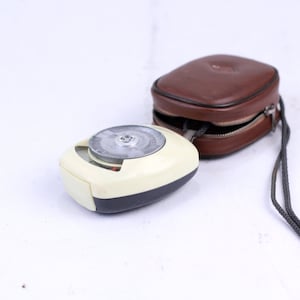 May include: A white and gray light meter with a brown leather case and a black strap. The light meter has a round dial with a needle.