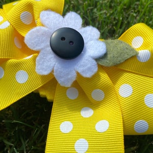 Puede incluir: Una cinta amarilla con lunares blancos y una flor de fieltro blanco con un botón negro en el centro. La flor tiene una hoja de fieltro verde.