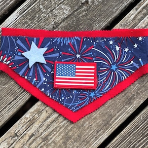 Puede incluir: Bandana patriótica para perros con fondo azul marino con fuegos artificiales y estrellas blancas y rojas. Una estrella azul claro y un parche de la bandera estadounidense están centrados en la bandana, que tiene un borde rojo.