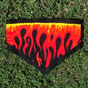 Puede incluir: Un bandana para perro de fieltro negro con un patrón de llamas rojas y amarillas. El bandana tiene un borde amarillo.