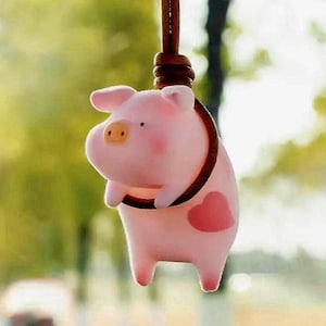 Puede incluir: Un amuleto de cerdo rosa con un corazón en un lado, colgando de un cordón marrón.