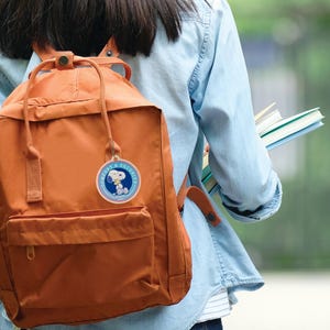May include: An orange backpack with a Snoopy tag that reads "Comfort & Security." The backpack has a front pocket and adjustable straps. A person is holding books and wearing a denim shirt.