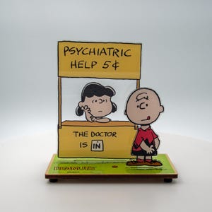 May include: A decorative Peanuts-themed figurine featuring Lucy in a psychiatric help booth with the text "PSYCHIATRIC HELP 5¢" and "THE DOCTOR IS IN." Charlie Brown stands nearby. The base is green with the word "PEANUTS".