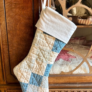 May include: A Christmas stocking made from quilted fabric featuring a patchwork pattern of light beige and faded blue squares. The stocking has a solid white fabric cuff at the top and a white fabric loop for hanging, with visible cross-hatch stitching.