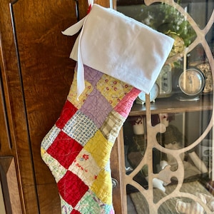 May include: A handmade Christmas stocking with a patchwork design. The stocking features a white cuff and a body composed of various fabric squares in red, yellow, pink, and blue. A white ribbon is attached for hanging. The stocking is hanging on a wooden cabinet.