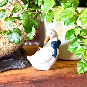 May include: A white ceramic goose figurine with a blue bow around its neck and a gold string for hanging. The goose has an orange beak and is set on a wooden surface, surrounded by greenery.