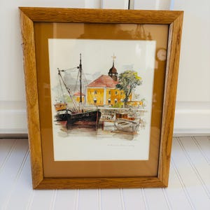 May include: A framed watercolor painting depicting a harbor scene with a large black fishing boat labeled SF 123, a smaller rowboat, and a yellow building with a red roof and a steeple. The artwork is enclosed in a light brown wooden frame.