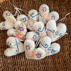 May include: A collection of heart-shaped ornaments with floral embroidery in shades of pink, blue, and orange. Each ornament has a white fabric base and a ribbon for hanging. The ornaments are arranged on a woven basket.
