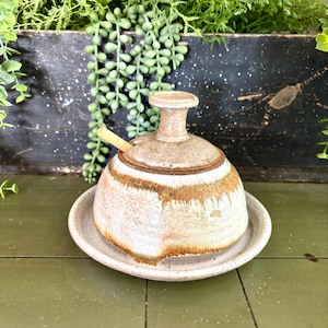 May include: A ceramic butter dish with a lid and a small spoon. The dish is off-white with brown and tan accents. It sits on a matching saucer. The background includes greenery and a dark wooden box.