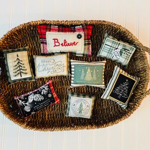 May include: A wicker basket filled with eight small, handmade, fabric Christmas pillows. The pillows feature various Christmas-themed designs, including a tree, "Merry Christmas", "Believe", and "Fresh Cut Christmas Trees".
