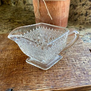 Peut inclure: Un pot à crème ou une saucière en verre transparent avec un motif décoratif en relief. Le pot à crème a une base carrée et une poignée.