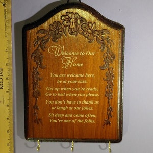 May include: A wooden key holder with a welcome home message and floral carvings. The key holder has five hooks for hanging keys. The text reads: "Welcome to Our Home. You are welcome here, be at your ease. Get up when you're ready, Go to bed when you please. You don't have to thank us or laugh at our jokes. Sit deep and come often, You're one of the folks."