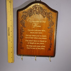 May include: A wooden key holder with a carved design of flowers and vines. It has a sign that reads "Welcome to Our Home" and a poem about being welcome in the home. The key holder has four hooks for hanging keys.