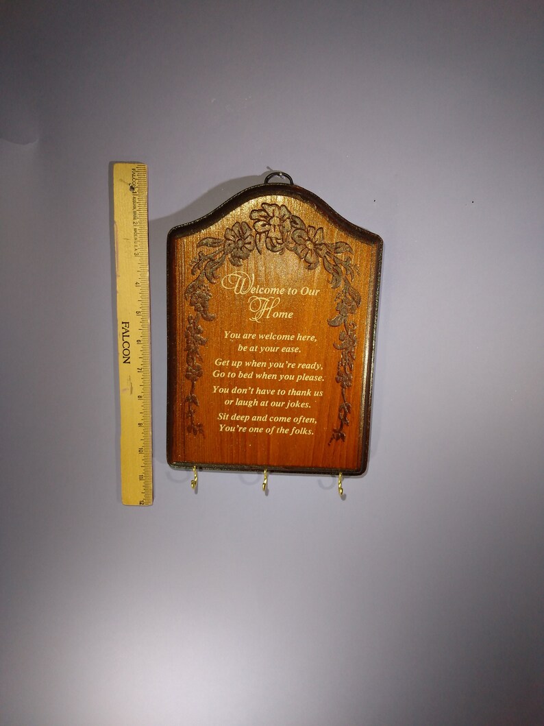 May include: A wooden key holder with a carved design of flowers and vines. It has a sign that reads "Welcome to Our Home" and a poem about being welcome in the home. The key holder has four hooks for hanging keys.