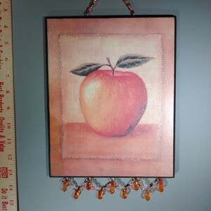 May include: A framed picture of a red apple with a brown wooden frame and a beaded orange and white garland hanging from the bottom.