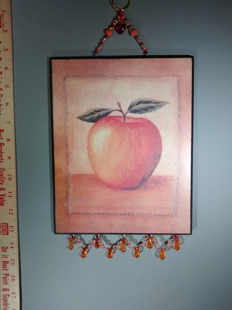 May include: A framed picture of a red apple with a brown wooden frame and a beaded orange and white garland hanging from the bottom.