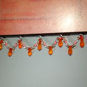 May include: A beaded garland with orange and clear glass beads. The garland has a scalloped shape and hangs from a wooden surface.