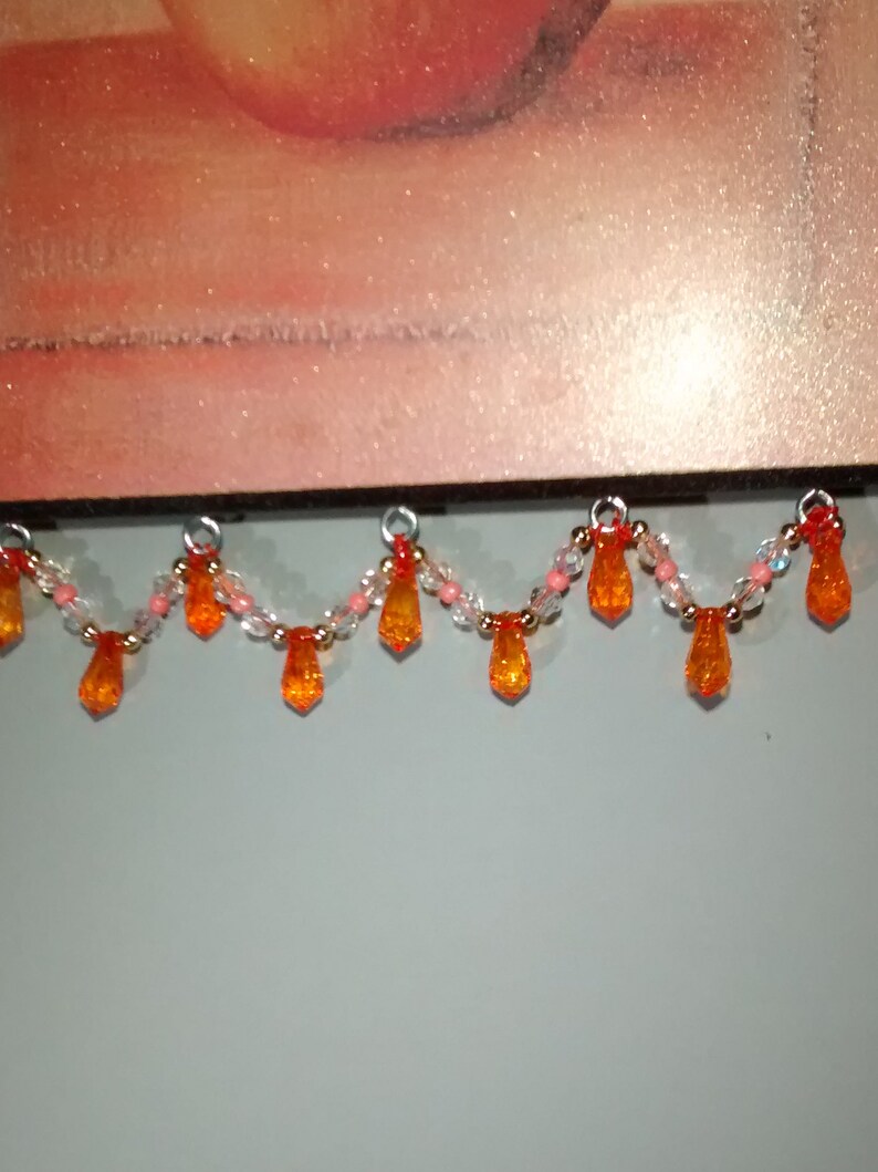 May include: A beaded garland with orange and clear glass beads. The garland has a scalloped shape and hangs from a wooden surface.
