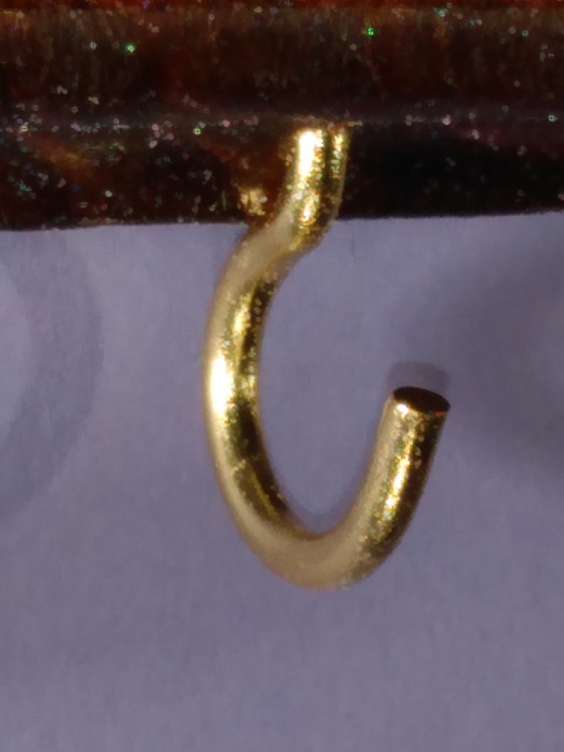 May include: A gold-colored metal hook with a rounded shape. The hook is designed to hang items on a surface.