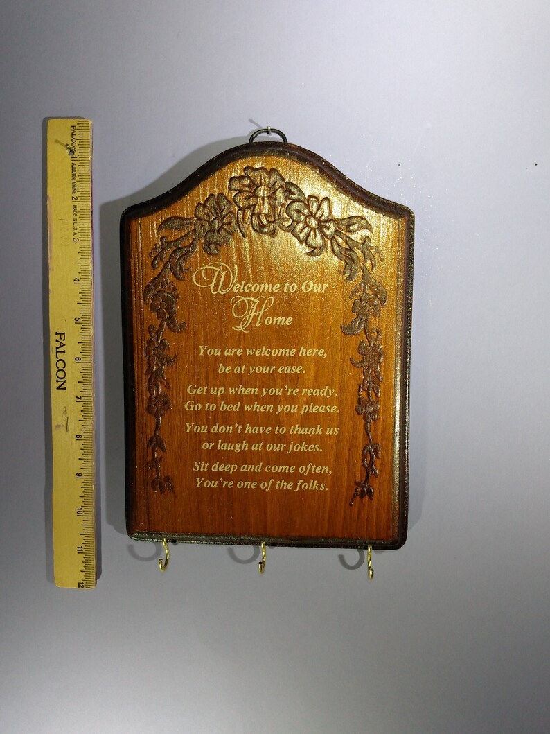 May include: Wooden sign with a gold inscription that reads "Welcome to Our Home". The sign has a carved floral design and three hooks on the bottom.