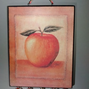 May include: A square wooden wall hanging featuring an illustration of a red apple, with a beaded fringe. The apple is set against a light pink background with a white border.