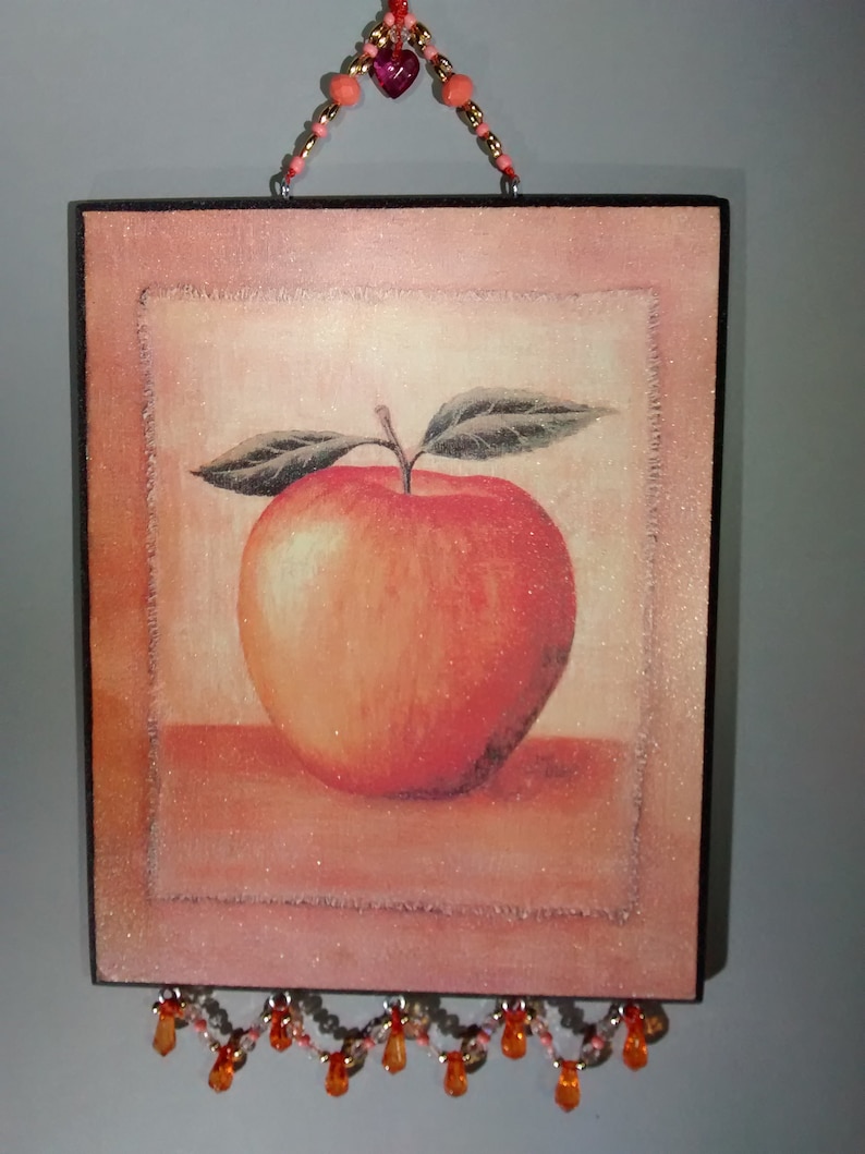 May include: A square wooden wall hanging featuring an illustration of a red apple, with a beaded fringe. The apple is set against a light pink background with a white border.